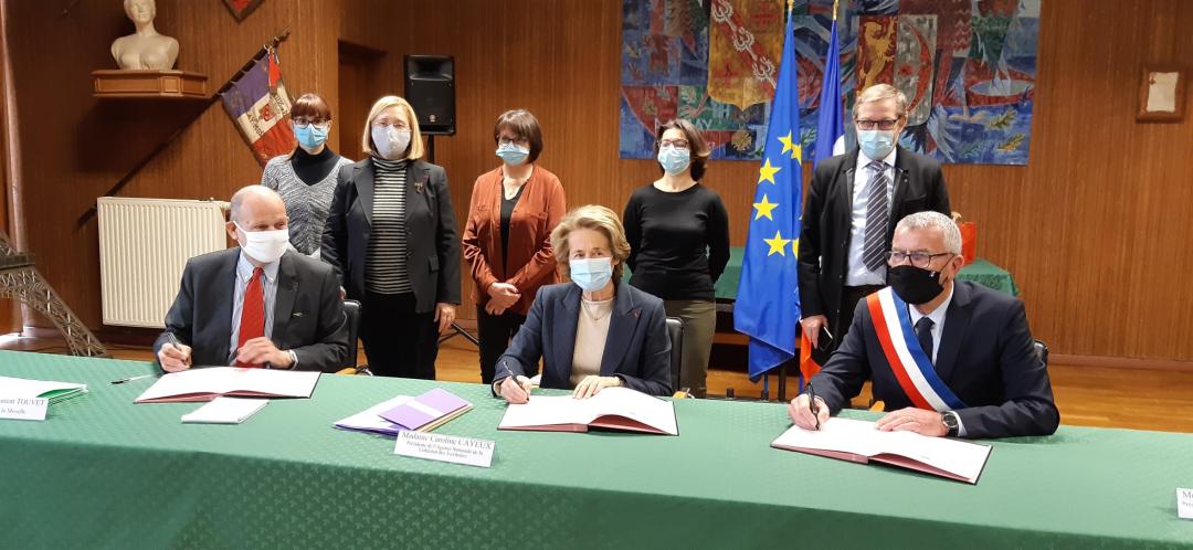 Trois personnes signent officiellement des parapheurs sur une talbe verte. L'un deux porte l'écharpe triclore des élus. Derrière eux, debouts, un groupe de collaborateurs. 