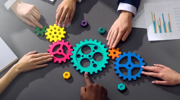 Photo illustrative représentant les mains de plusieurs personnes qui assemblent ensemble des rouages de couleur, posés sur une table.