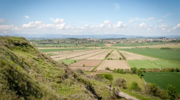 Payage avec au 1er plan un pente verdoyante, puis des champs agricoles et, en arrière-plan, un village dans un vallon et des collines au fond.