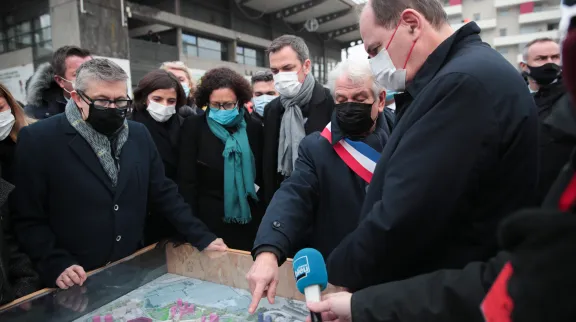 Jean Castex regarde une maquette en relief d'un quartier, présentée par le maire avec une écharpe tricolore, en extérieur et avec plein de gens autour de la maquette. 