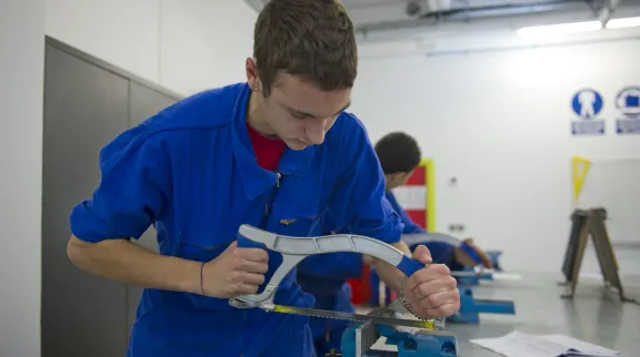 Jeune garçon en bleu de travail, en train de scier du métal sur un établi