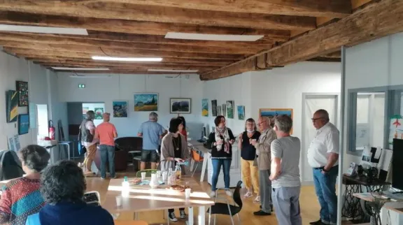 Dans une grande salle, avec des poutres anciennes au plafond, des personnes en petits groupes regardent une exposition de tableaux ou discutent dans un coin.