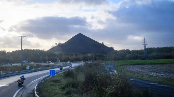 Vu sur la silhouette d'un terril, derrière une autoroute, avec le soleil couchant dans un ciel nuageux derrière le terril.