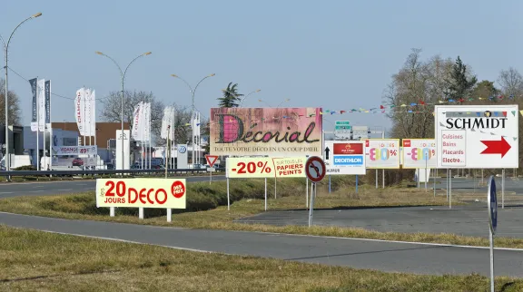 Vue d'une entrée de ville avec plein de panneaux publicitaires pour la zone commerciale, que l'on distingue en arrière plan.