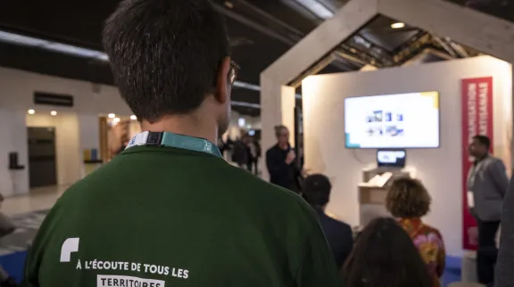 Vue de dos d'un homem en t-shirt vert marqué "A l'écoute de tous les territoires" qui regarde une conférence.