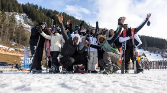 Photo : Un groupe de personnes en tenue de ski pose ensemble sur une piste enneigée, levant les bras avec enthousiasme. En arrière-plan, des chalets, des remontées mécaniques et une forêt de sapins sur une montagne enneigée.